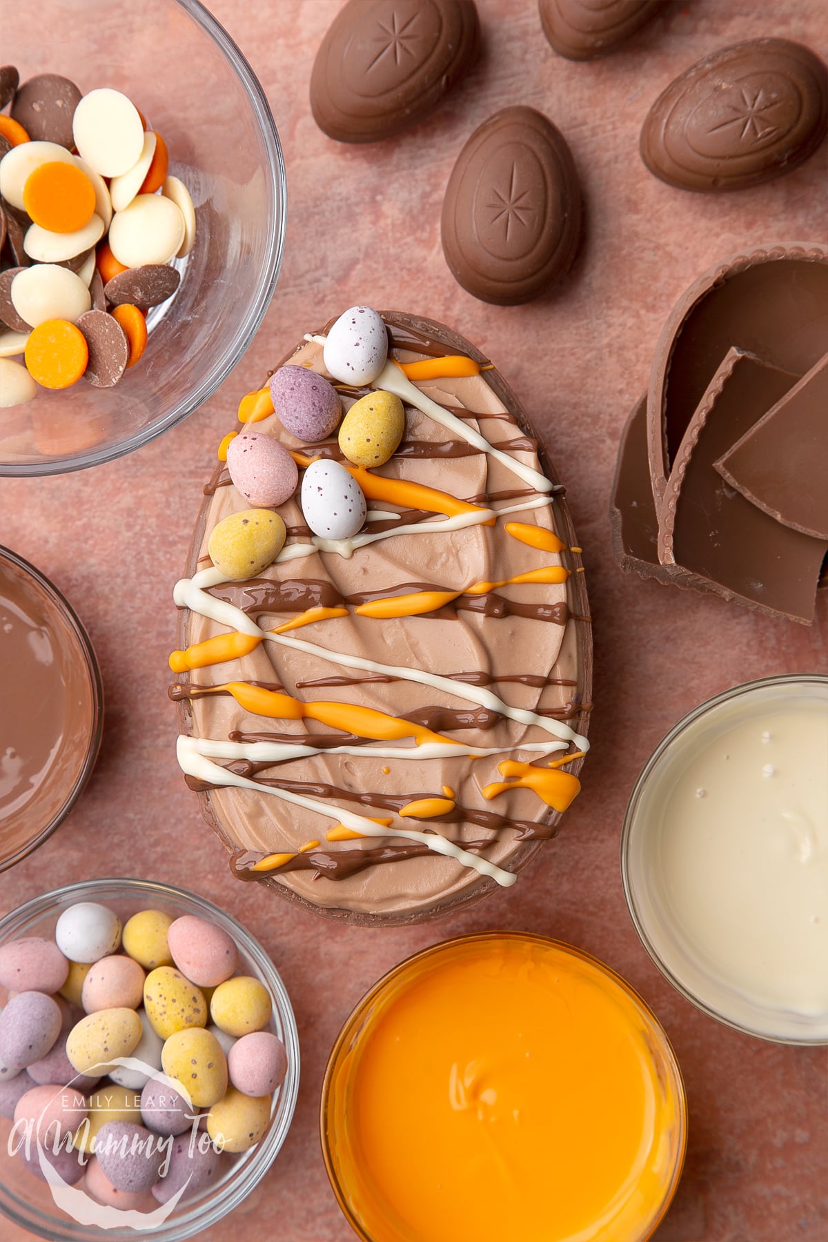 An Easter egg cheesecake on a small marble board. The chocolate cheesecake filling sits in a chocolate Easter egg shell, drizzled with white, milk and orange chocolate. The egg is surrounded by ingredients.