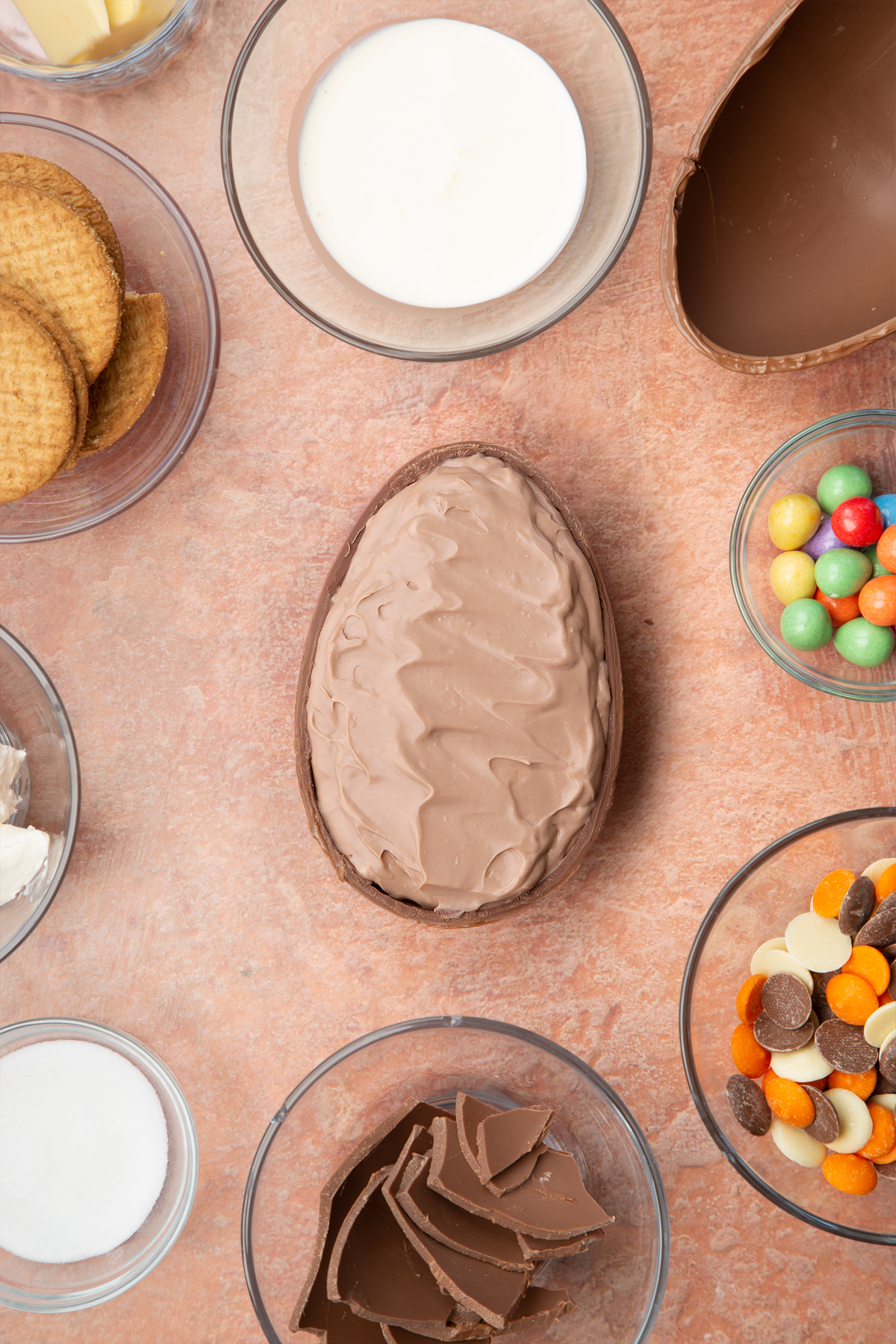 Half a chocolate Easter egg, cut side up, containing chocolate cheesecake filling. The egg is surrounded by ingredients to make Easter Egg cheesecake.