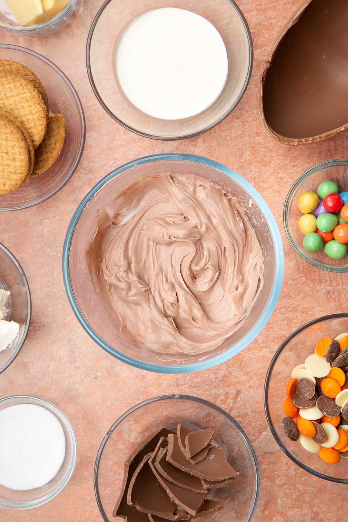 A glass bowl containing chocolate cheesecake filling. The bowl is surrounded by ingredients to make Easter Egg cheesecake.