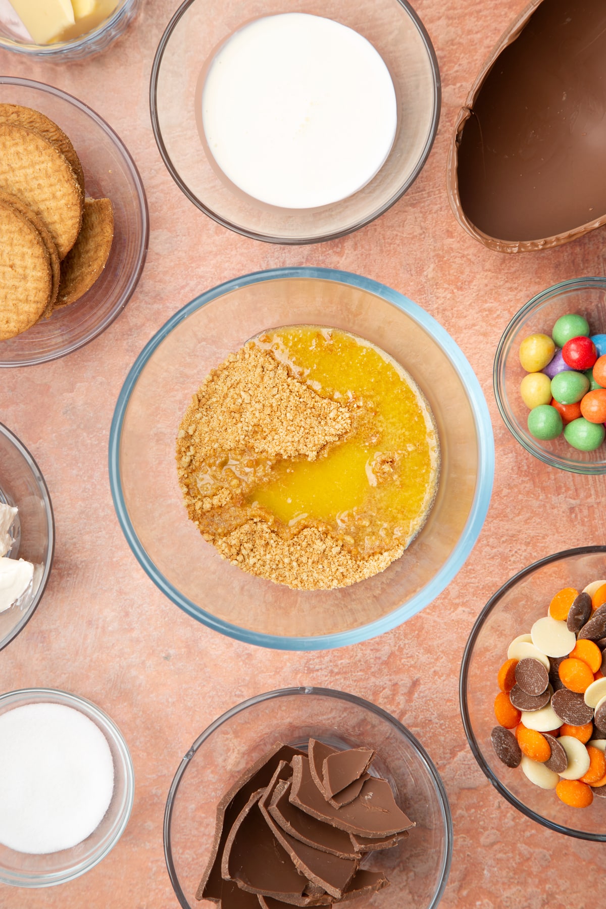 A glass bowl containing crushed digestive biscuits and melted butter. The bowl is surrounded by ingredients to make Easter Egg cheesecake.