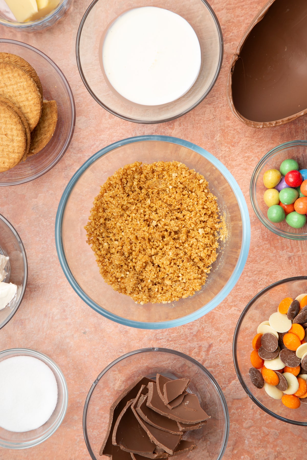 A glass bowl containing crushed digestive biscuits and melted butter mixed together. The bowl is surrounded by ingredients to make Easter Egg cheesecake.