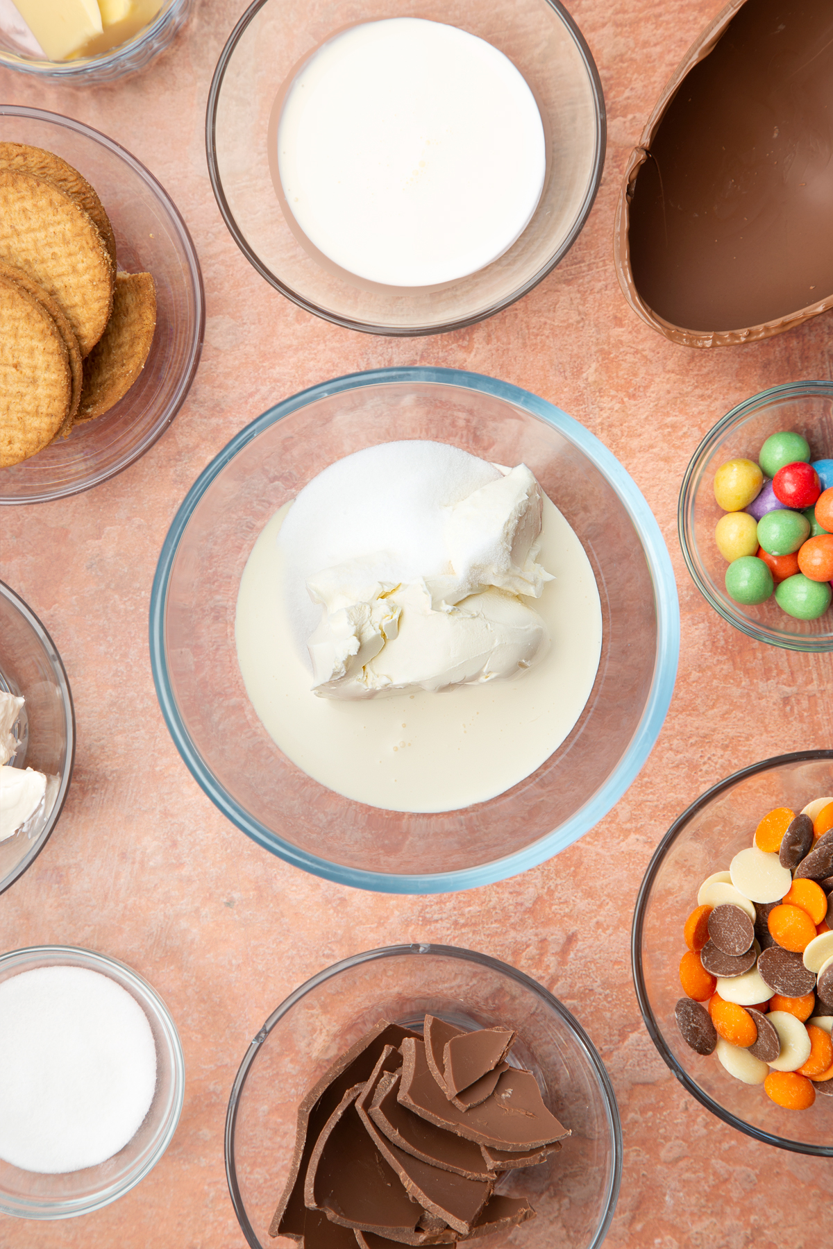 A glass bowl containing cream cheese, double cream and sugar. The bowl is surrounded by ingredients to make Easter Egg cheesecake.