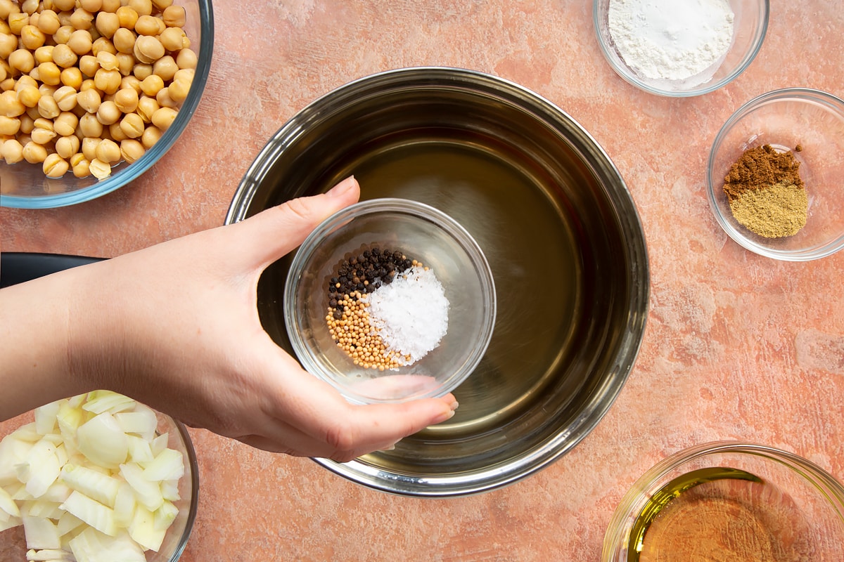 A hand holding a small bowl containing salt, mustard seeds and peppercorns over a pan of vinegar and water.