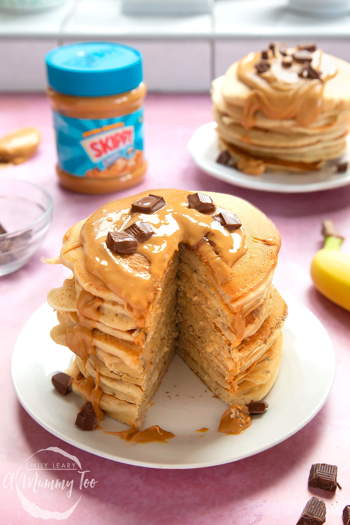 A stack of vegan peanut butter banana pancakes on a white plate. The stack is drizzled with more peanut butter and scattered with chunks of vegan chocolate. A large slice has been taken out of the whole stack, revealing the fluffy inner.