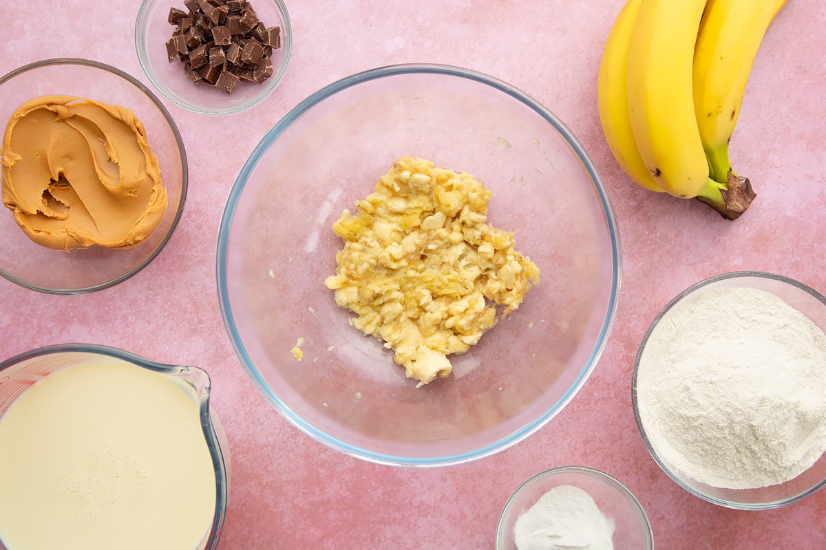 A bowl of mashed banana, surrounded by the ingredients for vegan peanut butter banana pancakes.