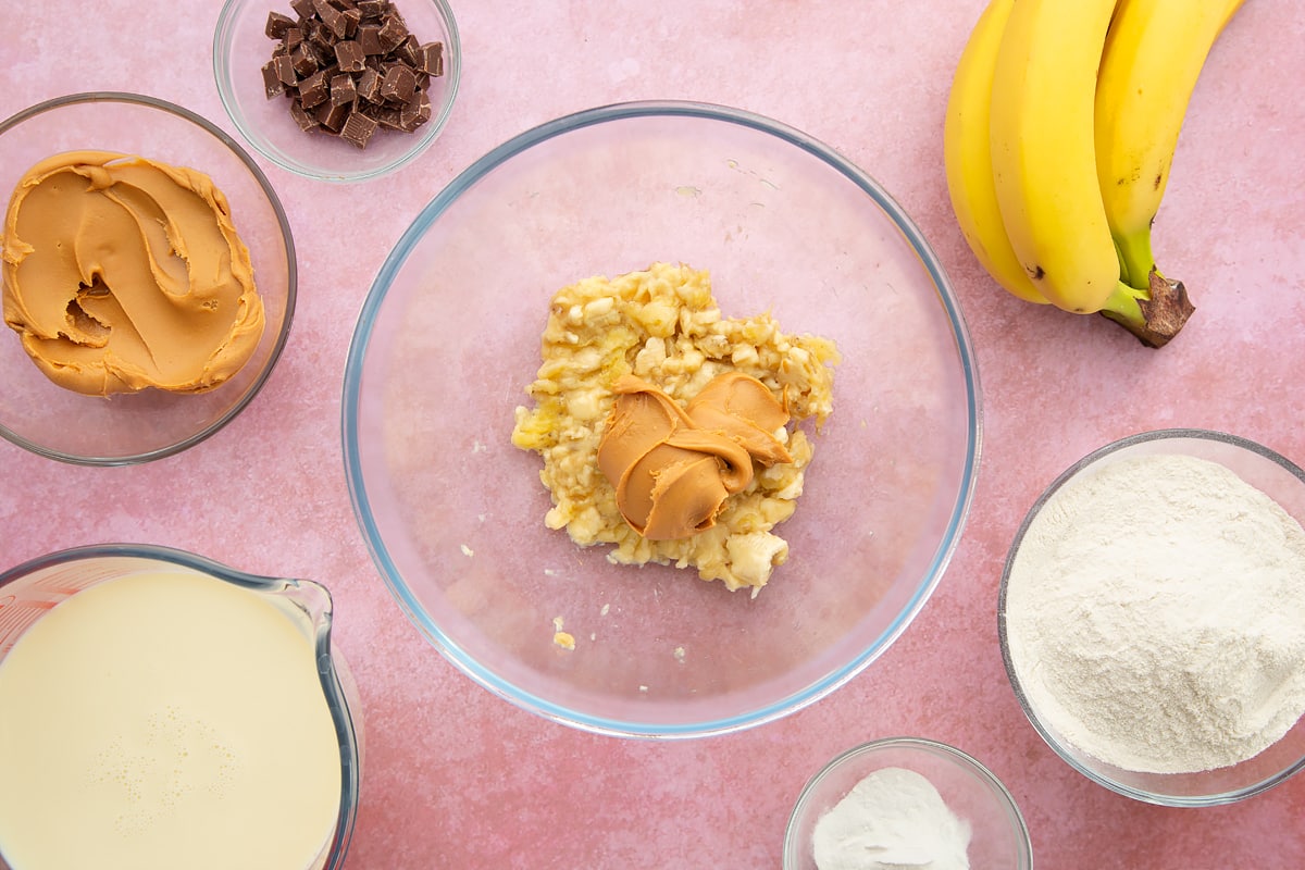 A bowl of mashed banana topped with smooth peanut butter, surrounded by the ingredients for vegan peanut butter banana pancakes.