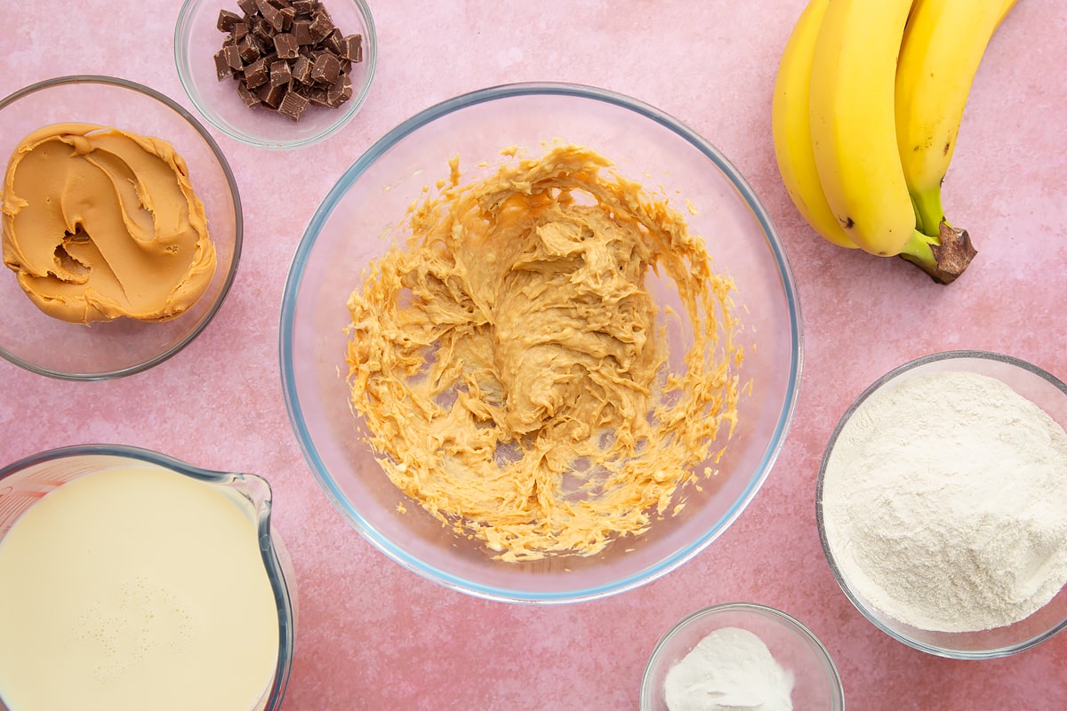 A bowl of banana and smooth peanut butter whisked together. The bowl is surrounded by the ingredients for vegan peanut butter banana pancakes.