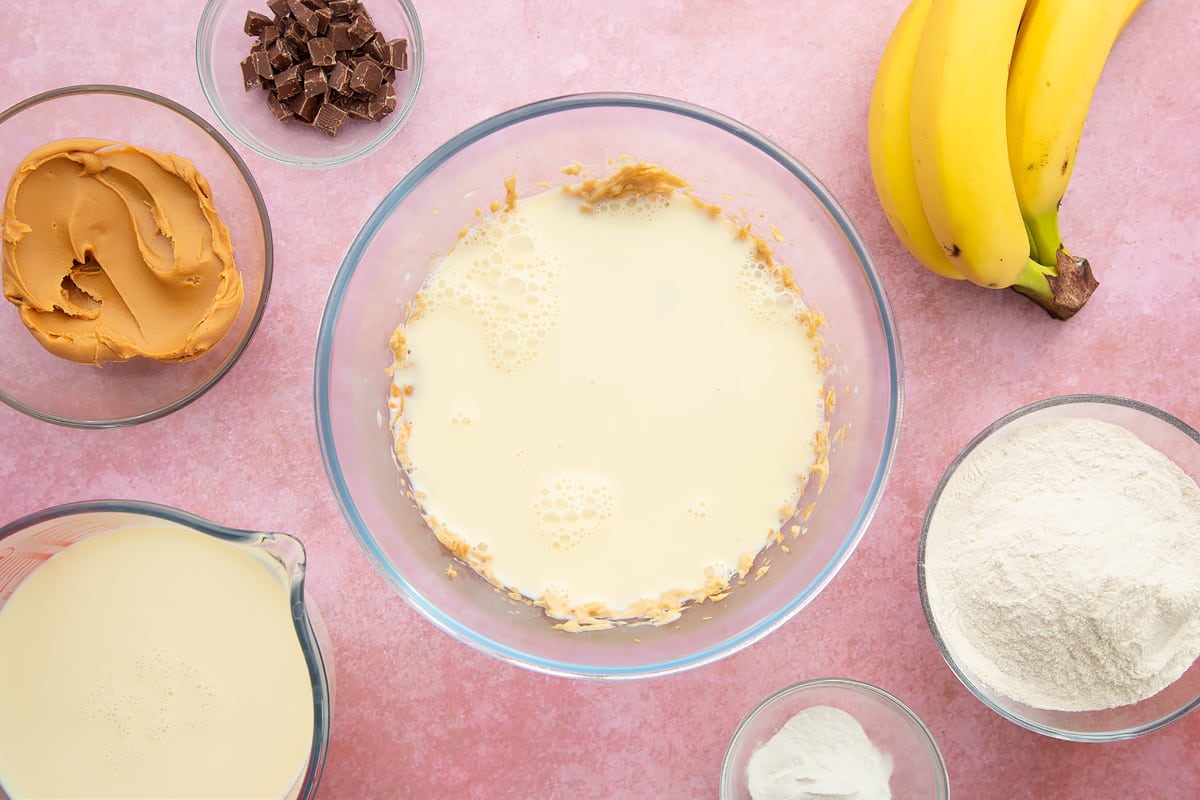 A bowl of banana and smooth peanut butter whisked together, topped with soya milk. The bowl is surrounded by the ingredients for vegan peanut butter banana pancakes.