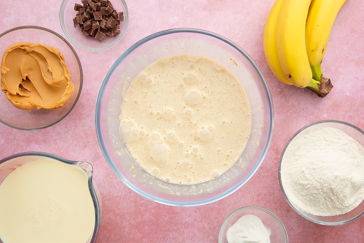 A bowl of banana, smooth peanut butter and soya milk whisked together. The bowl is surrounded by the ingredients for vegan peanut butter banana pancakes.