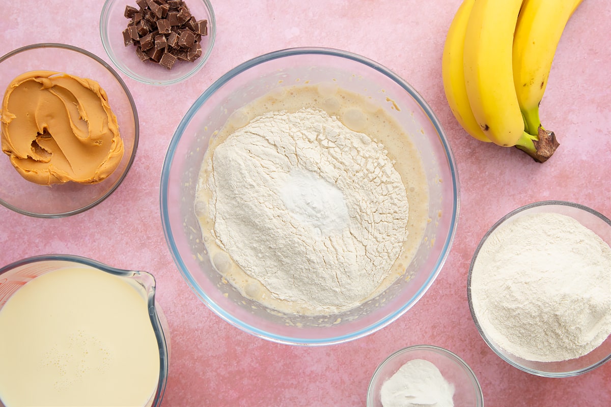 A bowl of banana, smooth peanut butter and soya milk whisked together, topped with flour and baking powder. The bowl is surrounded by the ingredients for vegan peanut butter banana pancakes.