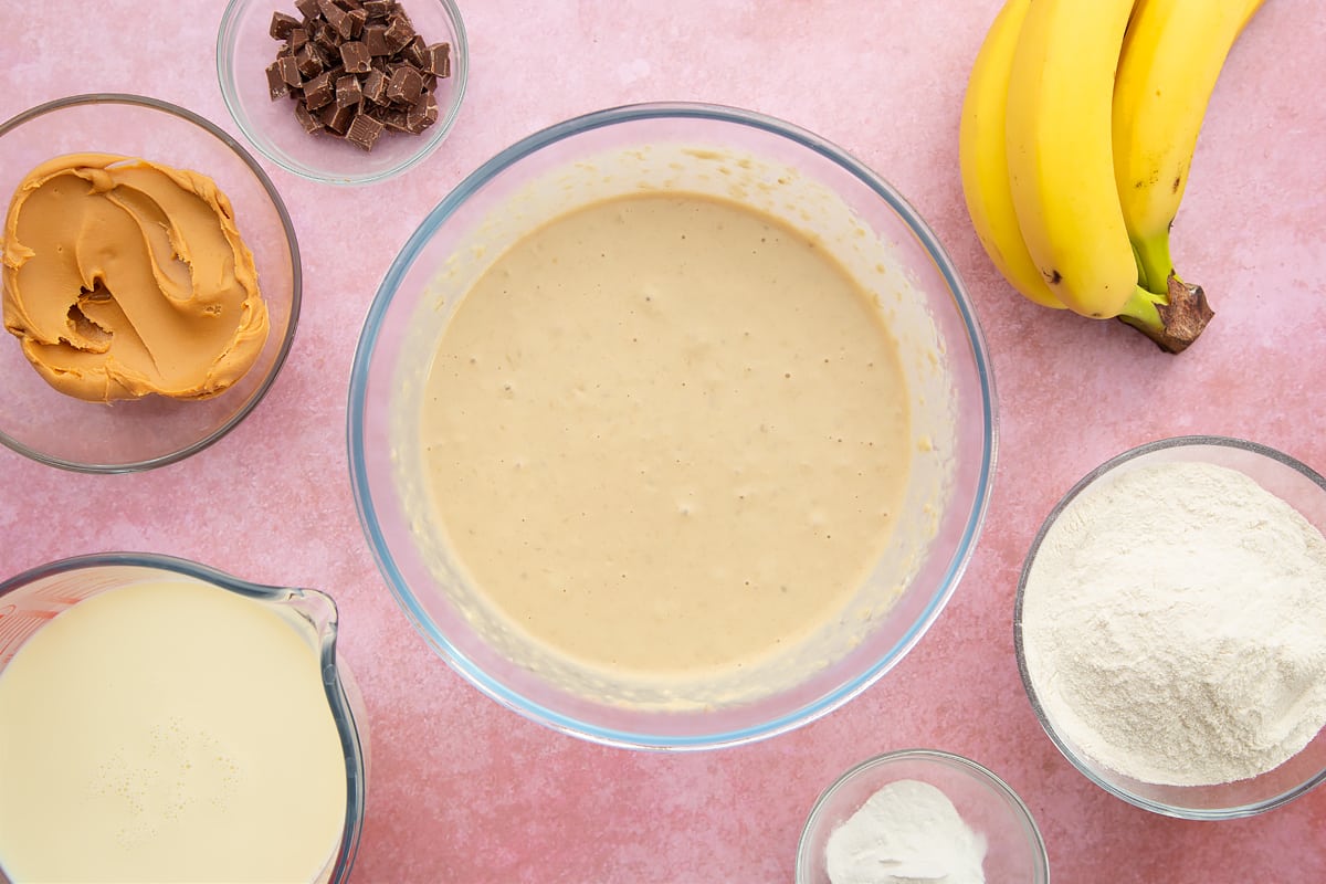 A bowl of banana, smooth peanut butter, soya milk, flour and baking powder whisked together. The bowl is surrounded by the ingredients for vegan peanut butter banana pancakes.