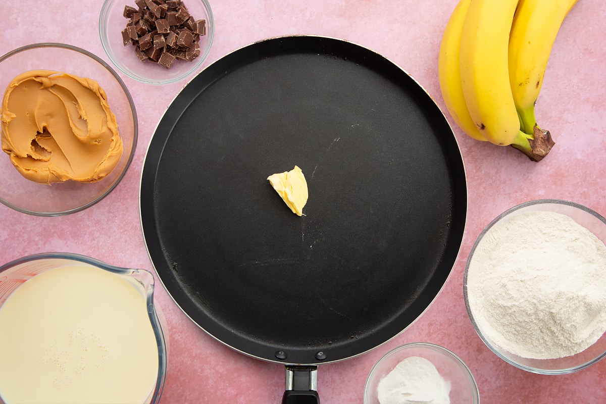 A frying pan with a knob of vegan spread in the centre. The pan is surrounded by the ingredients for vegan peanut butter banana pancakes.