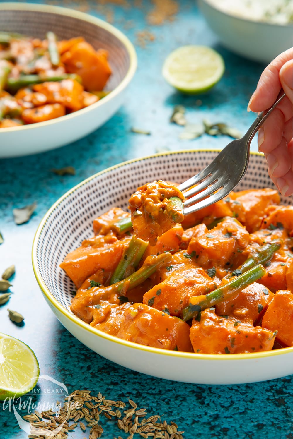 Vegan Sri Lankan curry in two yellow and black bowls. Ingredients are scattered around it. A fork lifts some a piece of sweet potato from the bowl in the foreground.