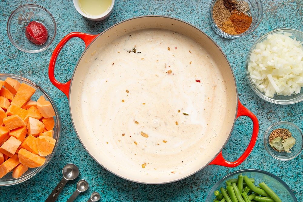 A large, shallow pan with coconut milk, tomato paste, fried onion, ginger, garlic, chilli and spices. The pan is surrounded by ingredients for vegan Sri Lankan curry.