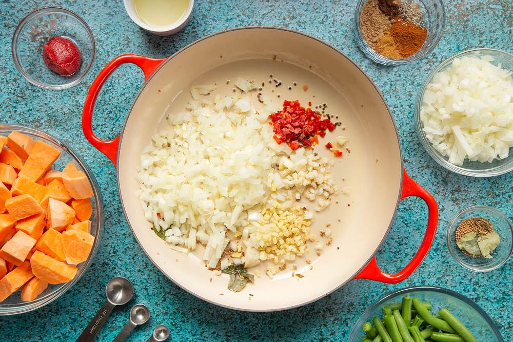 A large, shallow pan with mustard seeds, curry leaves and fennel seeds, onion, ginger, garlic and chilli frying in it. The pan is surrounded by ingredients for vegan Sri Lankan curry.