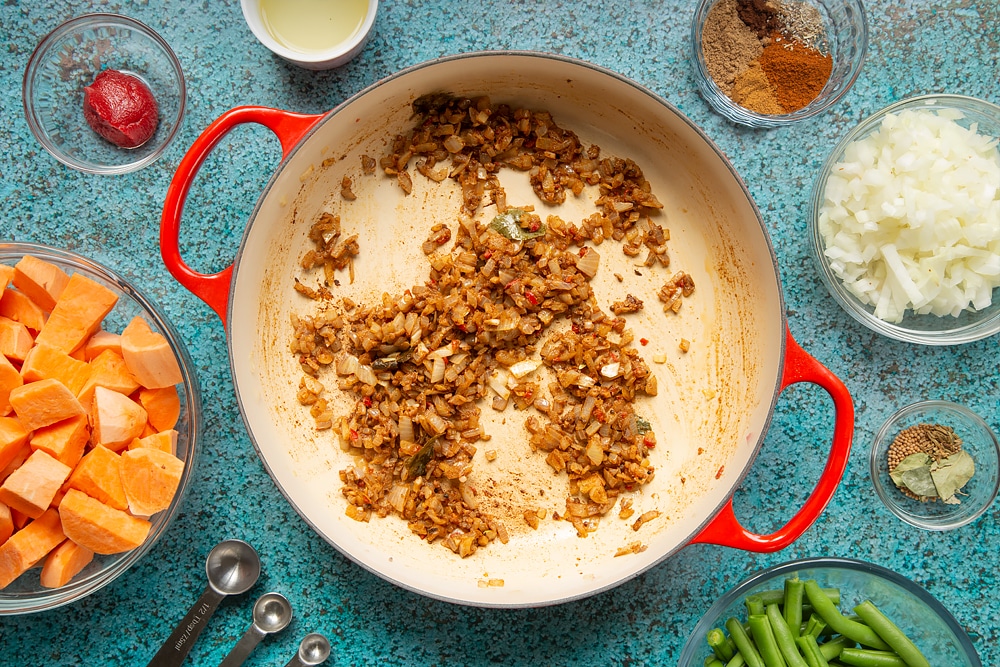 A large, shallow pan with onion, ginger, garlic, chilli and spices frying in it to create a deep brown mix. The pan is surrounded by ingredients for vegan Sri Lankan curry.