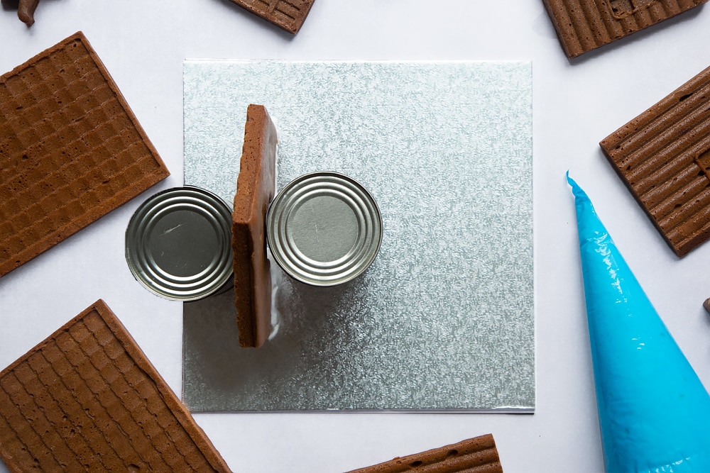 A silver cake board with a piece of gingerbread house wall standing on it. Two cans support the wall on either side.