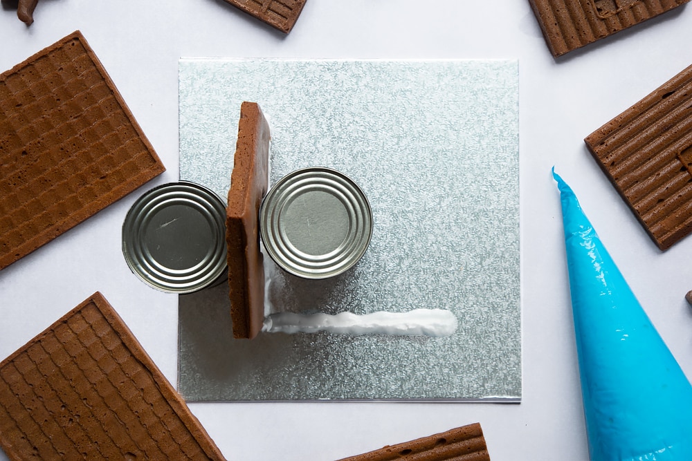 A silver cake board with a piece of gingerbread house wall standing on it. Two cans support the wall on either side. A further line of royal icing is piped where the next wall will be.