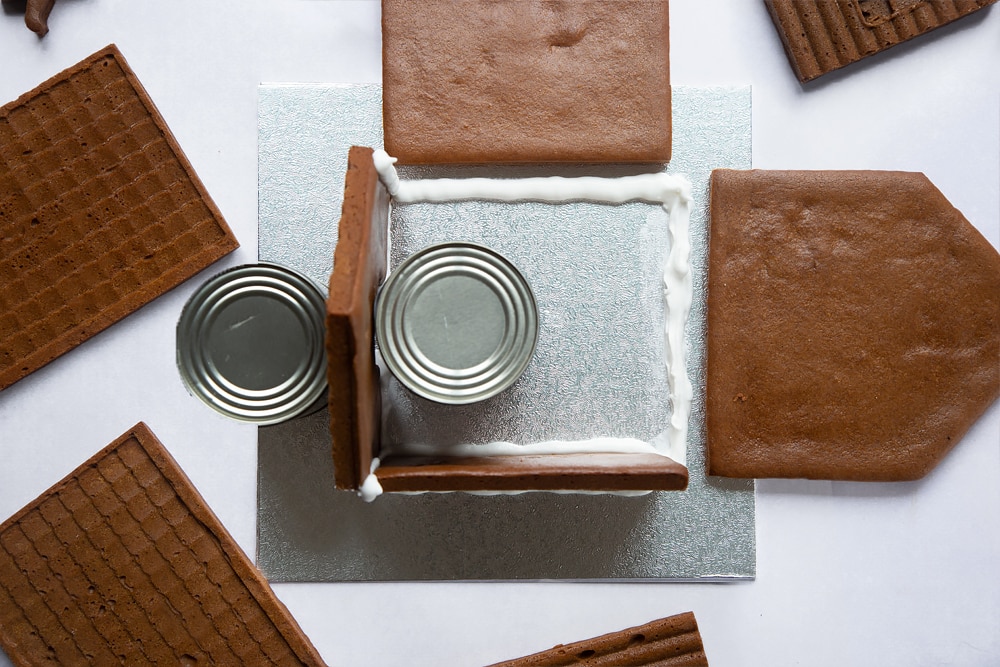 A silver cake board with two pieces of gingerbread house wall standing on it. Two cans support one wall on either side. A further line of royal icing is piped where the finals wall will be. they are lying either side of the board.