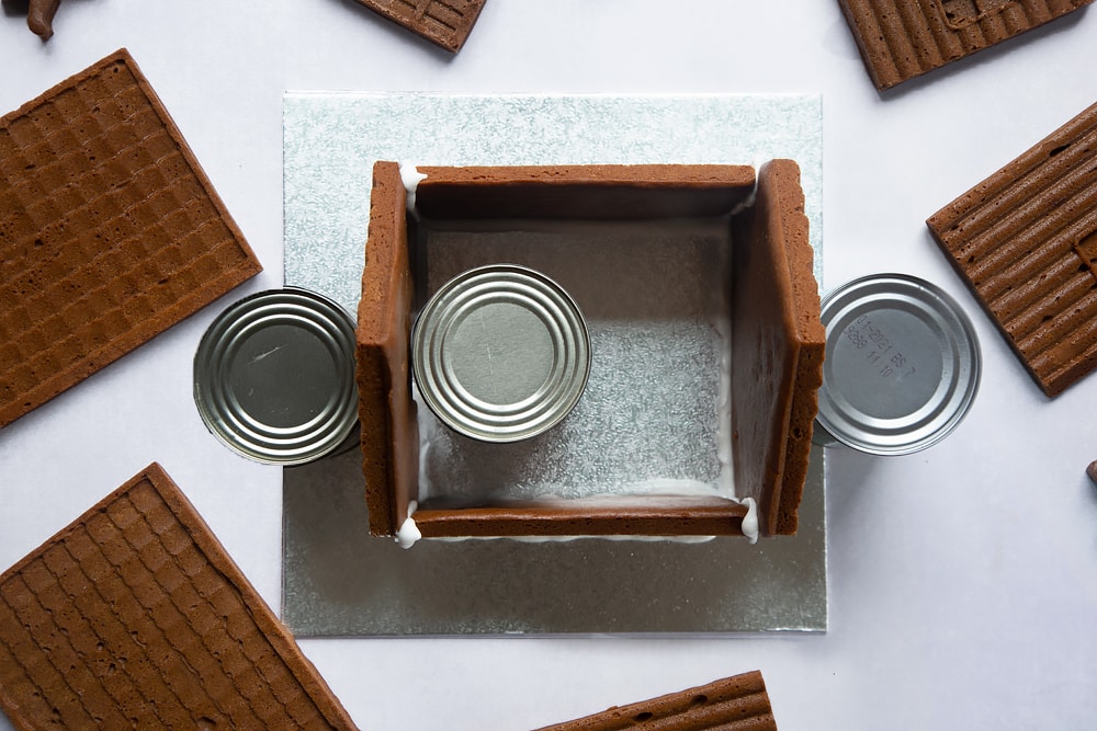 A silver cake board with all four walls of a standing on it. Three cans support the walls.