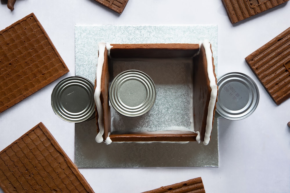 A silver cake board with all four walls of a standing on it. Three cans support the walls. Royal icing is piped along the top where the roof will be added.