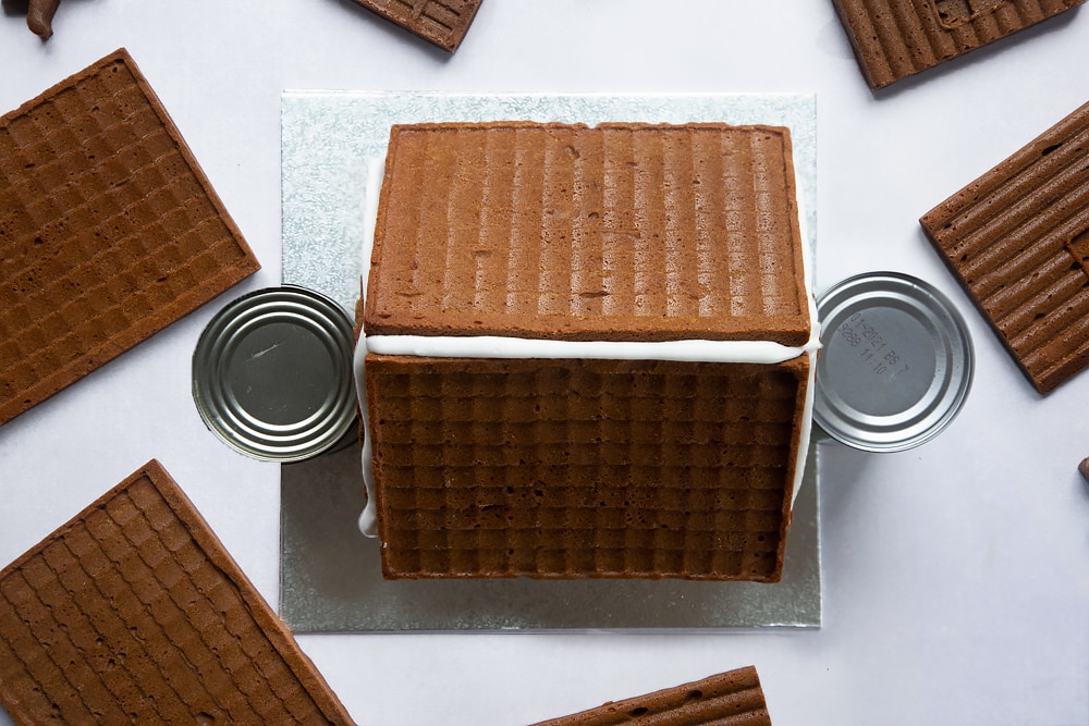 A silver cake board with all four walls of a standing on it and the two roof pieces on top. A line of royal icing is piped along the top where the two roof pieces meet. Two cans support the walls.