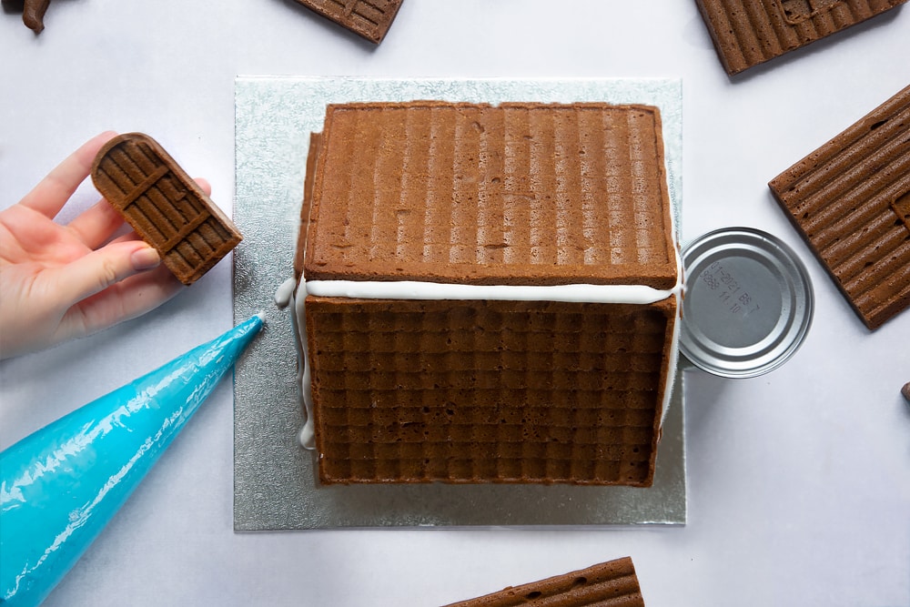 A silver cake board with a gingerbread house on top. A hand hold the gingerbread door.