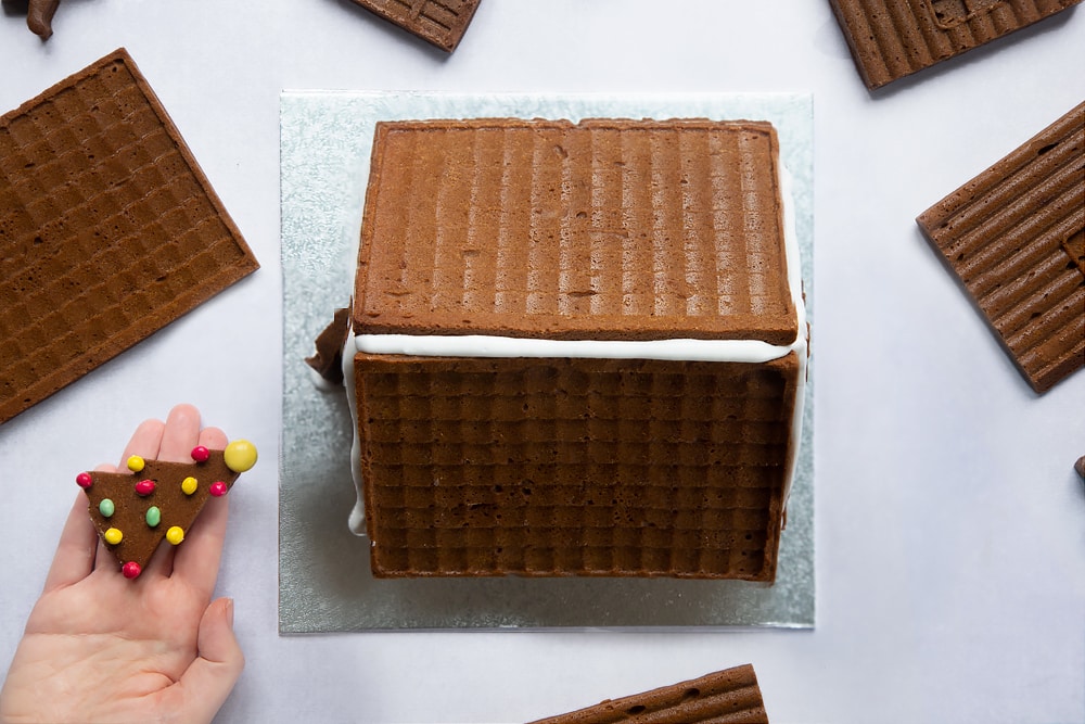 A silver cake board with a gingerbread house on top. A hand holds a decorated gingerbread Christmas tree, decorated with mini chocolate beans.