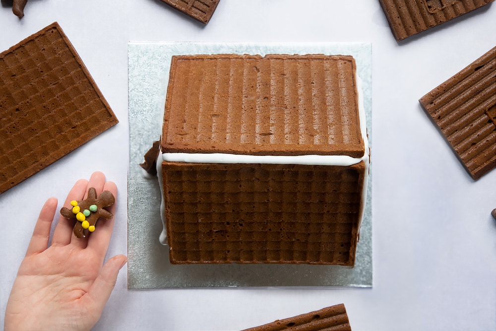 A silver cake board with a gingerbread house on top. A hand holds a decorated gingerbread person, decorated with mini chocolate beans.