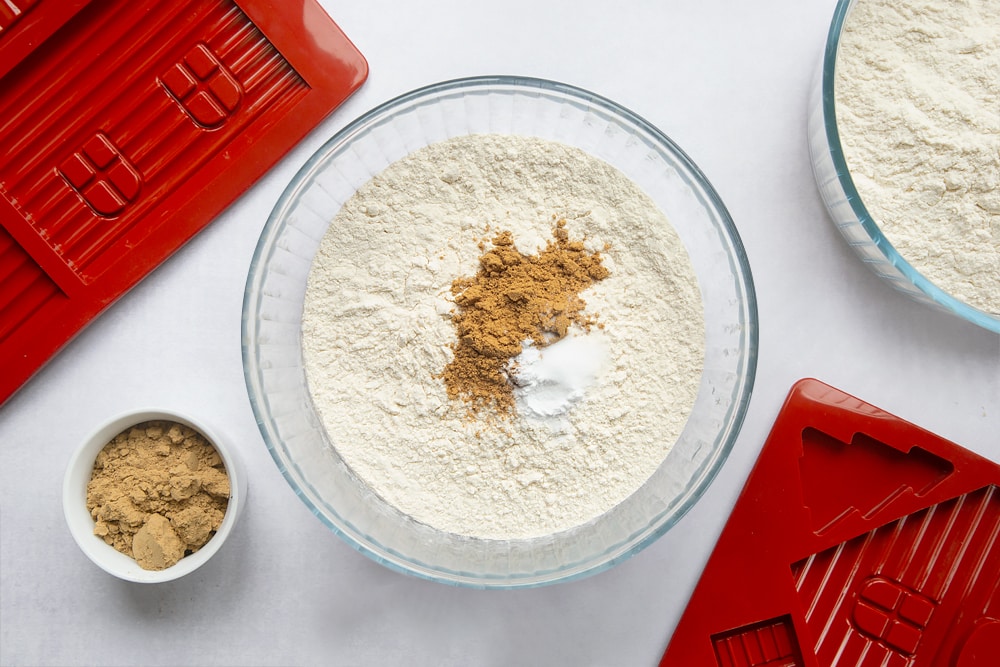 A bowl, surrounded by equipment to make a gingerbread house. In the bowl is flour, ground ginger and bicarbonate of soda. 