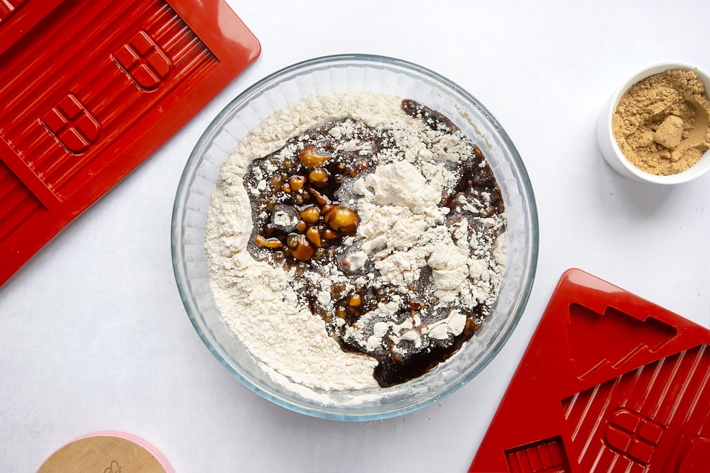 A bowl, surrounded by equipment to make a gingerbread house. In the bowl is flour mixed with ground ginger and bicarbonate of soda. Add to this is a syrup of golden syrup, black treacle, butter and sugar.