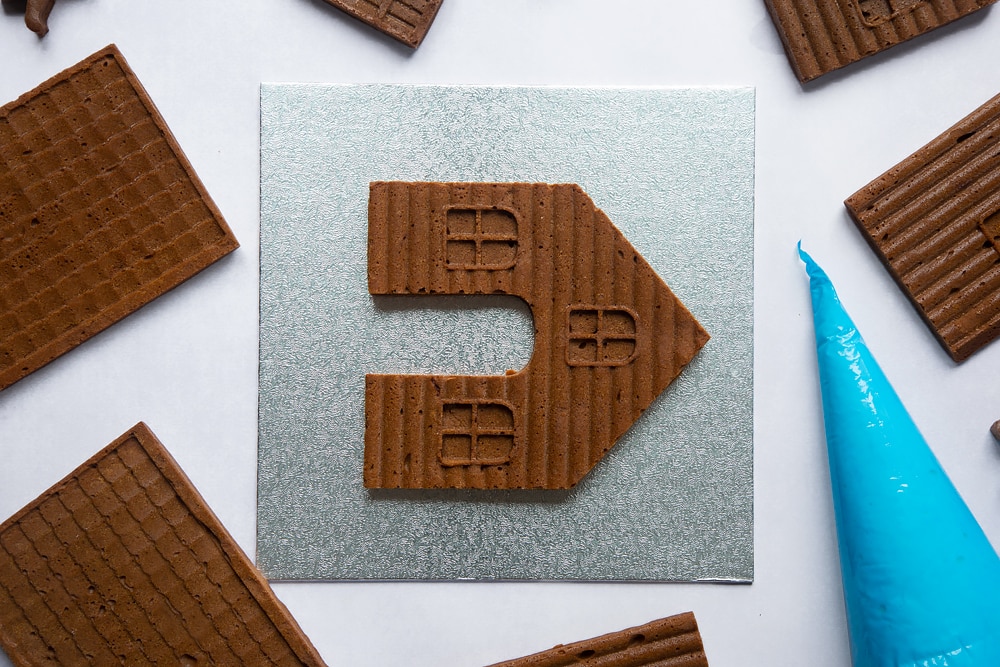 A silver cake board with a piece of gingerbread house on it. Other pieces of gingerbread house surround it.