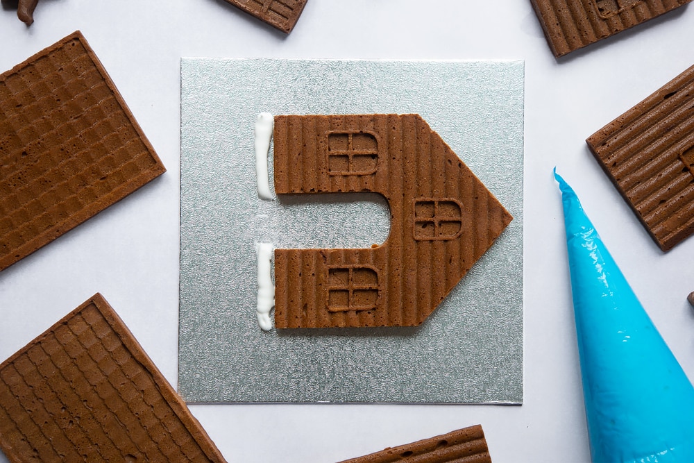 A silver cake board with a piece of gingerbread house on it. Royal icing piped on the board beside it.