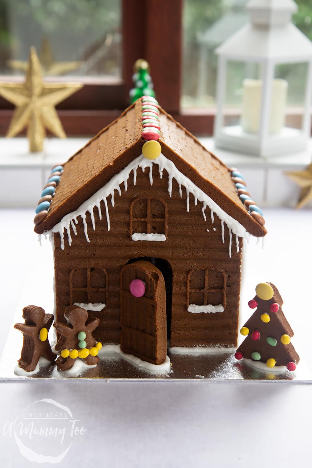 A detailed gingerbread house on a silver board. The house is decorated with smarties and mini chocolate beans. A gingerbread Christmas tree is shown to the left of the house, while two gingerbread people stand to the right.