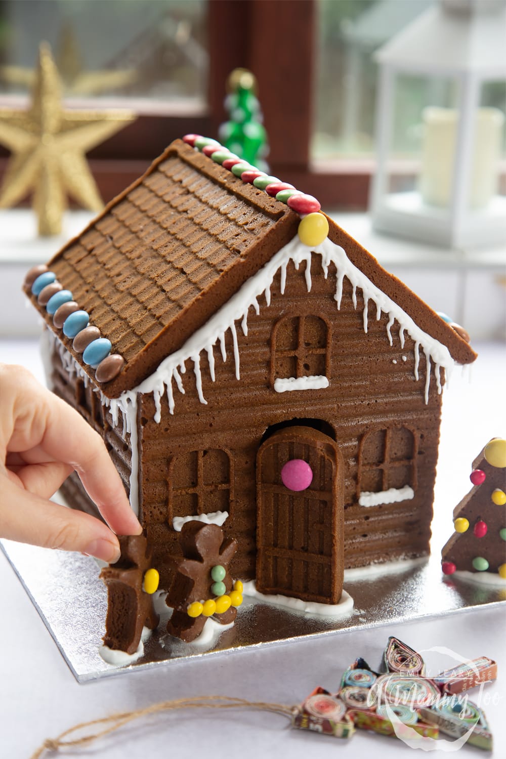 A detailed gingerbread house on a silver board. A hand is reaching in, positioning two gingerbread people in place on the board.