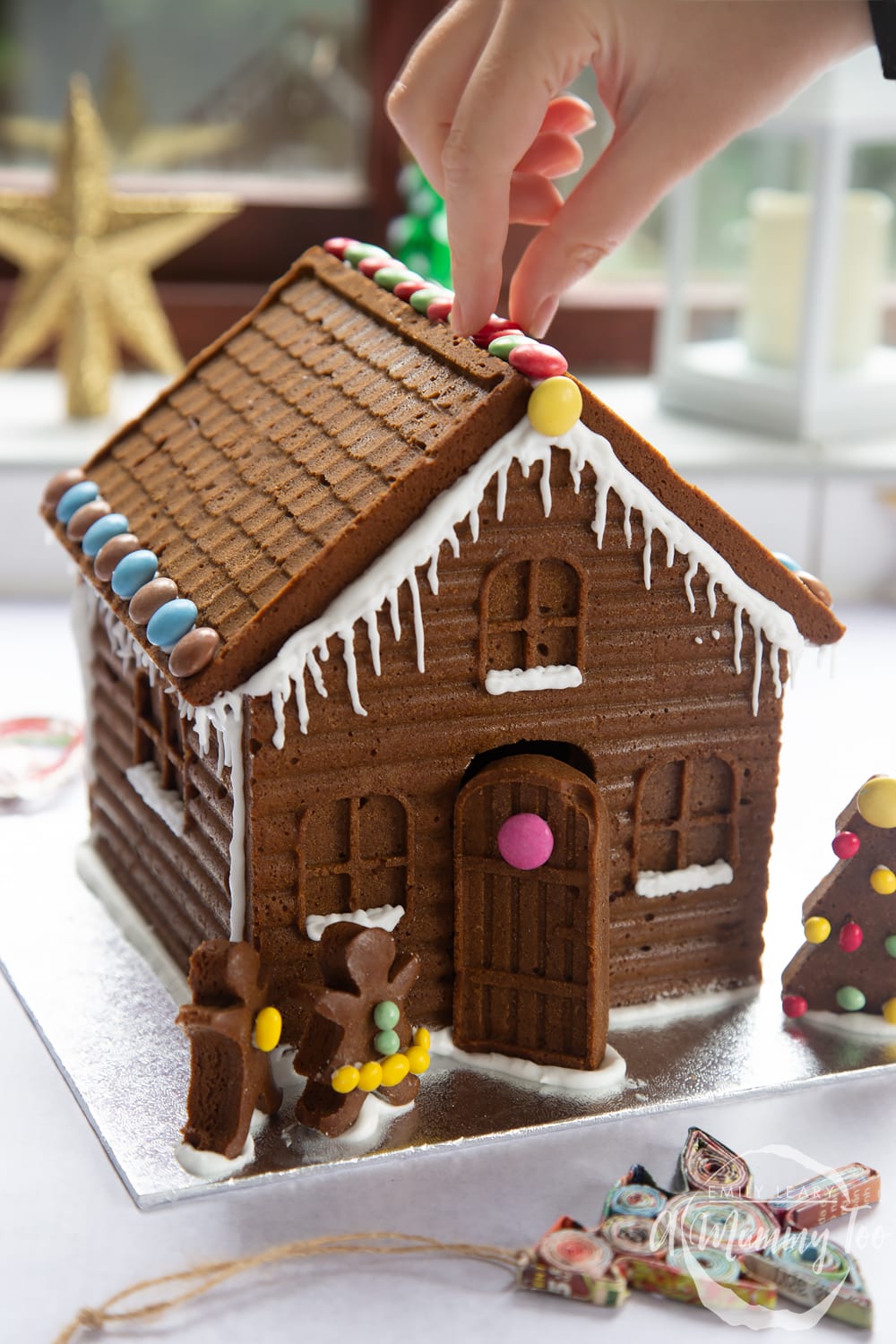 A detailed gingerbread house on a silver board. A hand is reaching in, positioning chocolate beans on the roof of the house.