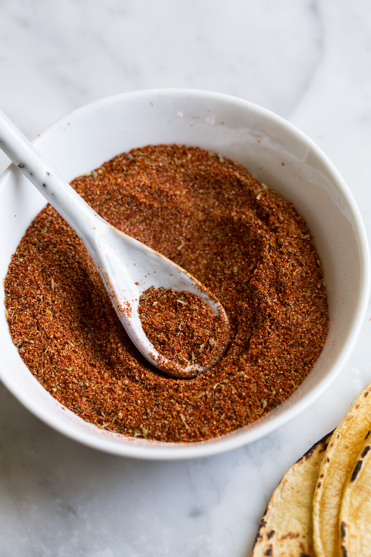 Blended taco seasoning in a small white ceramic bowl with a white ceramic spoon scooping up the seasoning.