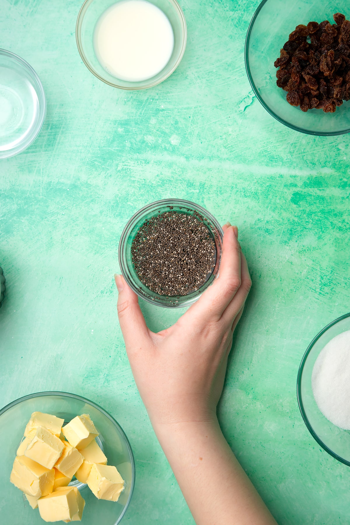 A hand holds a small bowl containing chia seeds mixed with water. Ingredients to make vegan Welsh cakes surround the bowl.