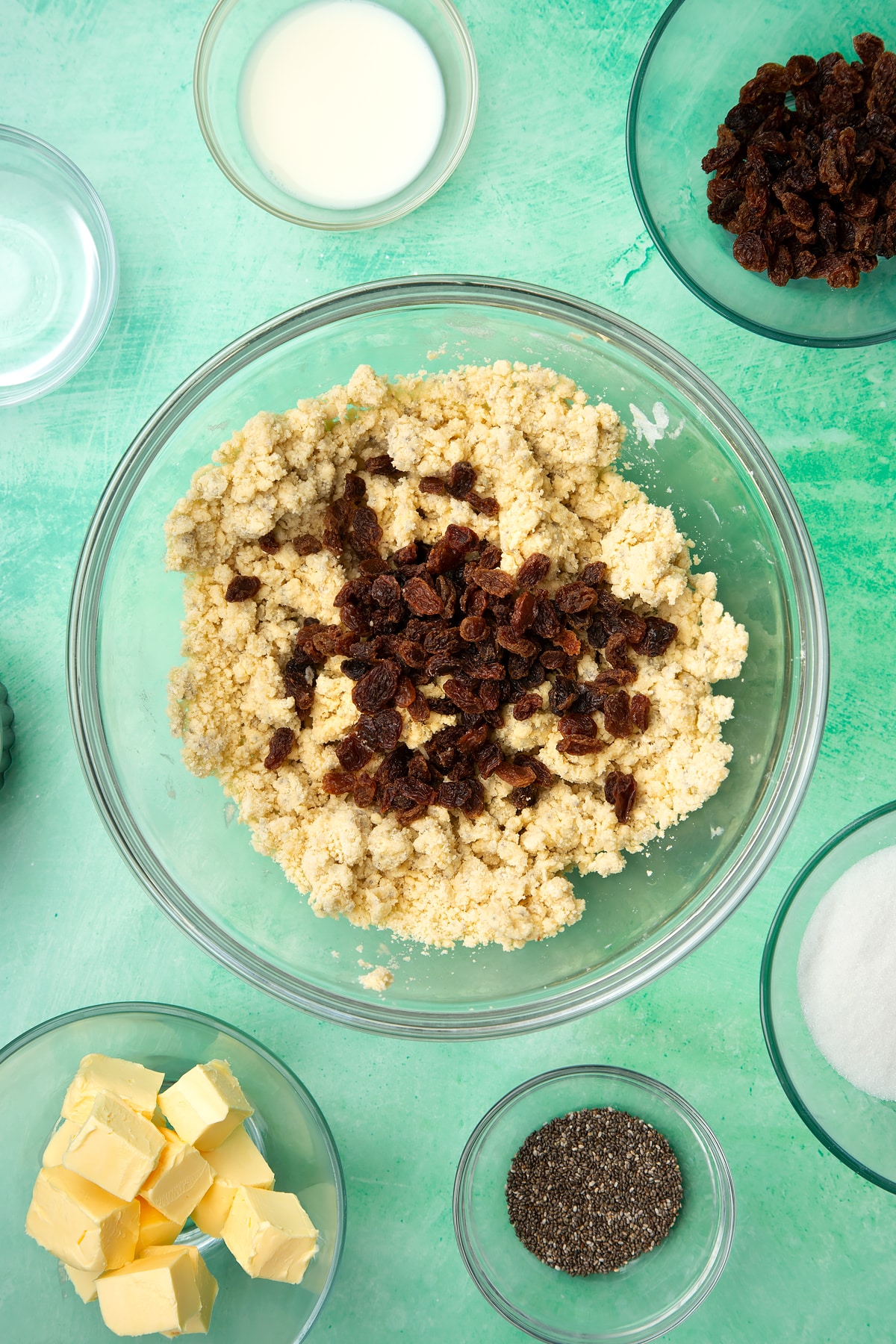 A glass mixing bowl containing self-raising flour, vegan margarine, chia seeds, sugar and plant milk mixed together, with sultanas on top. Ingredients to make vegan Welsh cakes surround the bowl.