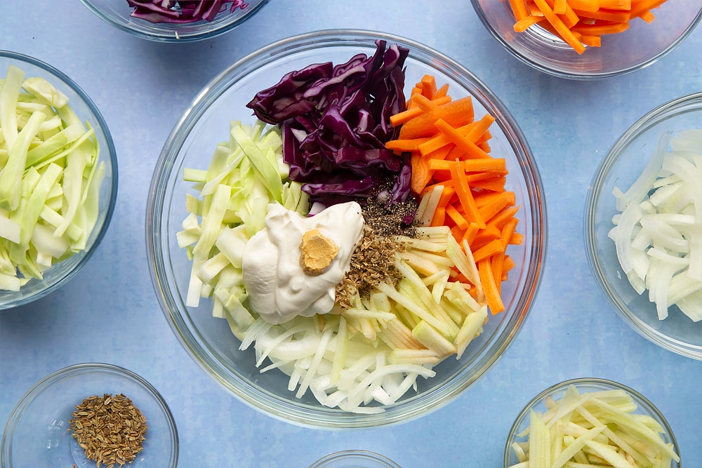 A glass mixing bowl containing carrot, onion, apple, white cabbage and red cabbage all cut into thin batons. Crème fraiche, mustard and fennel seeds are shown on top. Ingredients to make coleslaw without mayo surround the bowl.