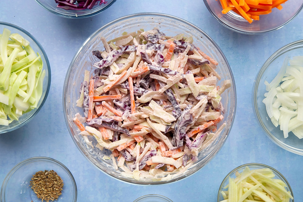 A glass mixing bowl containing shredded vegetables and creme fraiche. Ingredients to make coleslaw without mayo surround the bowl.