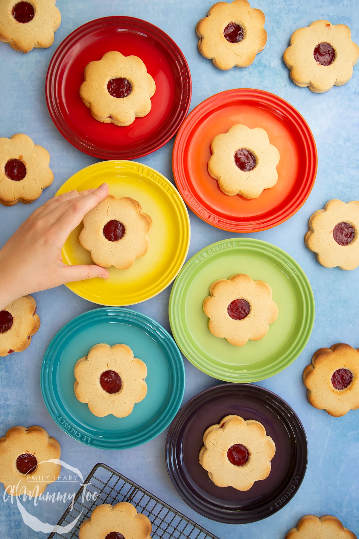 Giant jammie dodgers arranged on a variety of coloured small plates. A hand reaches in to take one from the yellow plate.