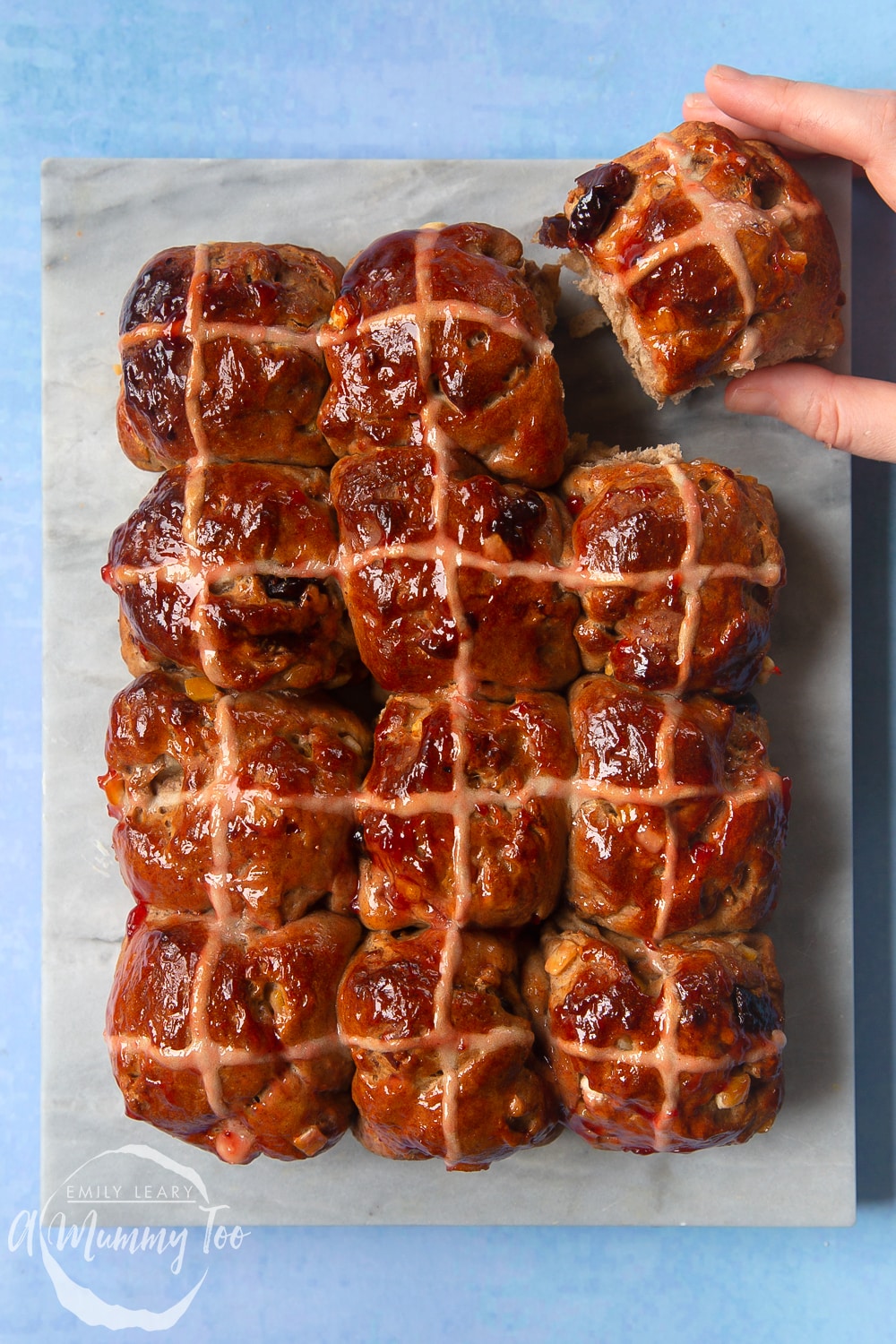 12 vegan hot cross buns on a marble board. A hand reaches in to take one.