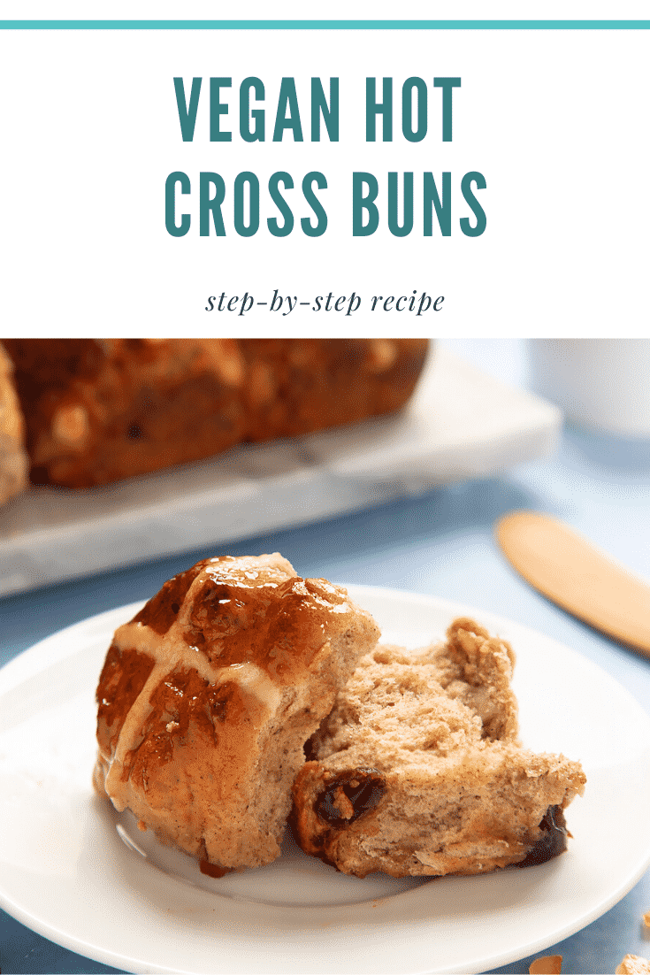 A vegan hot cross bun cut open on a white plate. More vegan hot cross buns are shown on a marble board in the background. A caption reads: vegan hot cross buns - step-by-step recipe