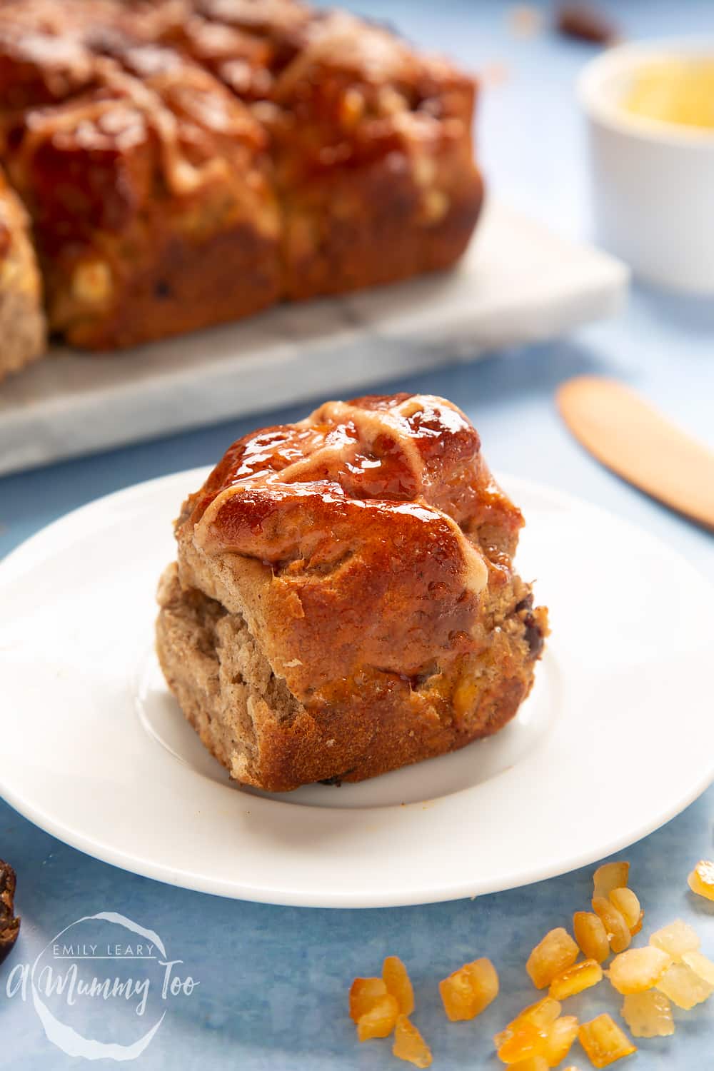 A vegan hot cross bun on a white plate. More vegan hot cross buns are shown on a marble board in the background.