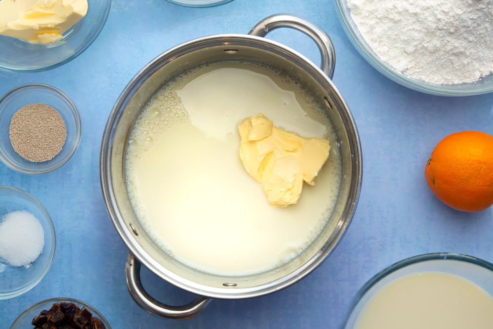 A saucepan containing plant milk and vegan margarine. The pan is surrounded by the ingredients to make vegan hot cross buns.