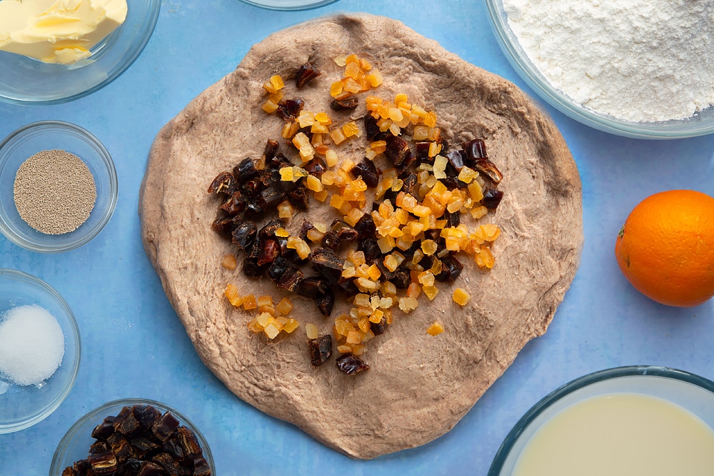 Vegan hot cross buns dough, spread out into a disc and topped with mixed peel and chopped dates. The dough is surrounded by vegan hot cross bun ingredients.