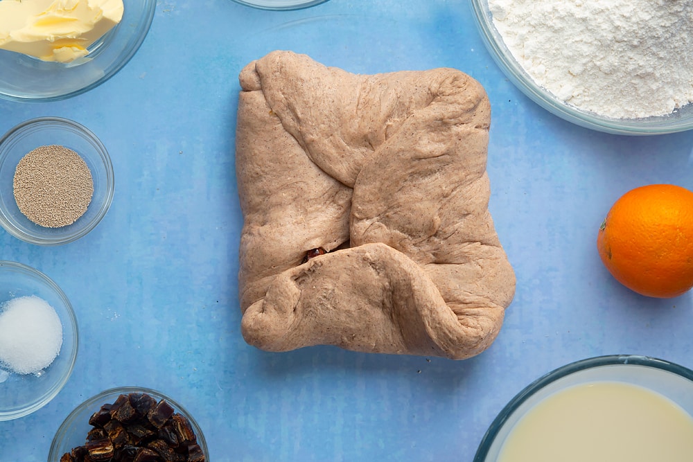 Vegan hot cross buns dough with mixed peel and chopped dates, folded into an envelope. The dough is surrounded by vegan hot cross bun ingredients.