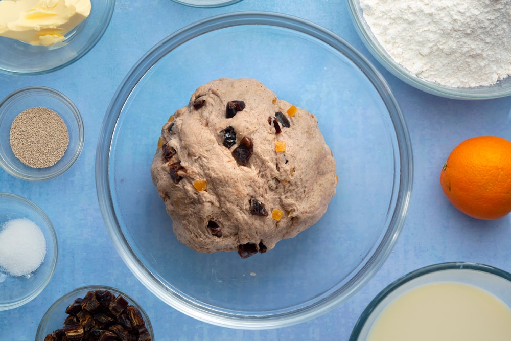 Vegan hot cross buns dough with mixed peel and chopped dates in a bowl. The dough is surrounded by vegan hot cross bun ingredients.