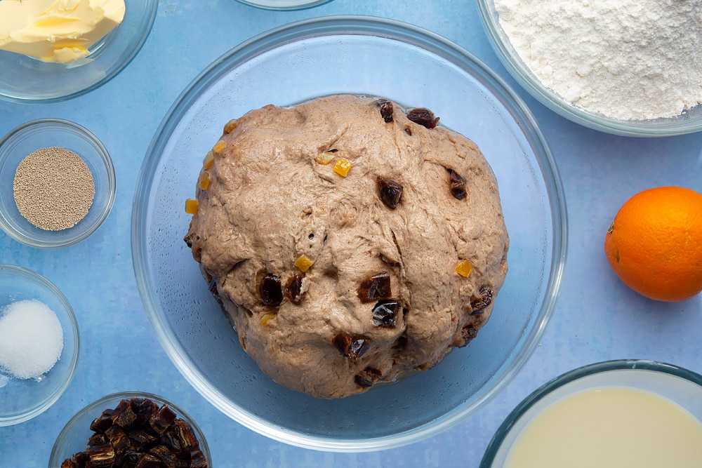 Vegan hot cross buns dough with mixed peel and chopped dates in a bowl, freshly proved. The dough is surrounded by vegan hot cross bun ingredients.